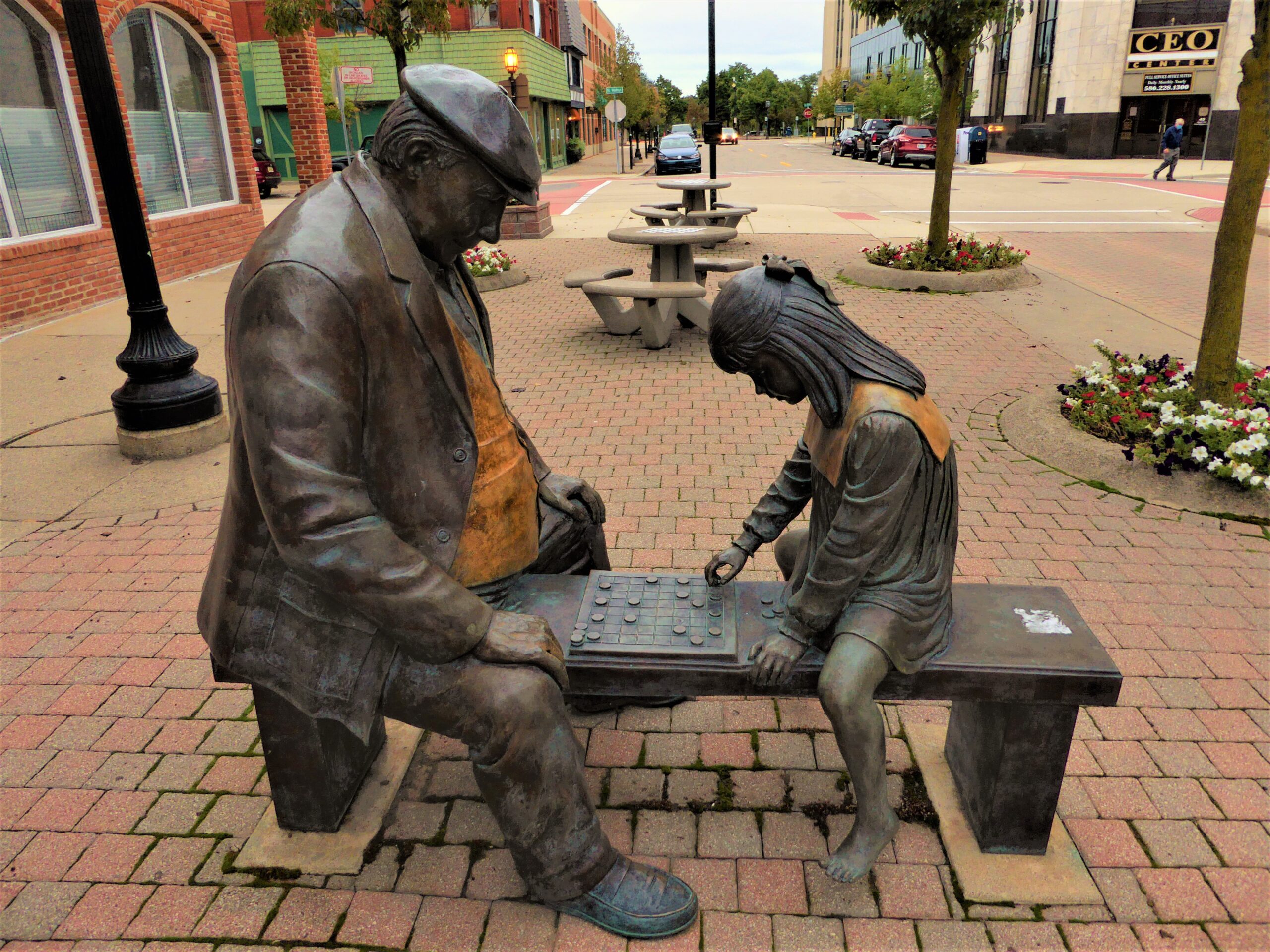 Photo of a sculpture of an older man playing checkers with a young girl, both are sitting on a bench.