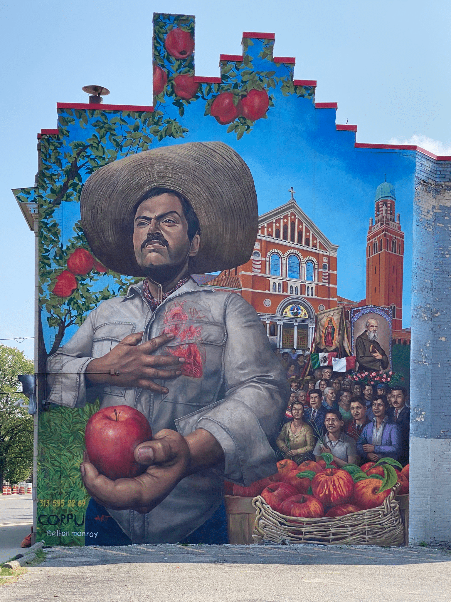 Photo of a mural of a Latinx man holding an apple, holding his hand to his heart, behind him there are portraits of community members and a church in Southwest Detroit